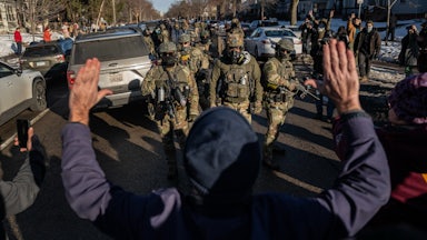 A protester stand with their hands raised above their head as masked federal immigration agents walk towards them