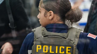 An ICE agent stands with her back to the camera and looks to the side