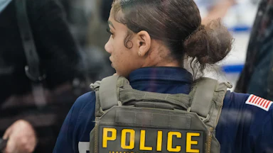 An ICE agent stands with her back to the camera and looks to the side