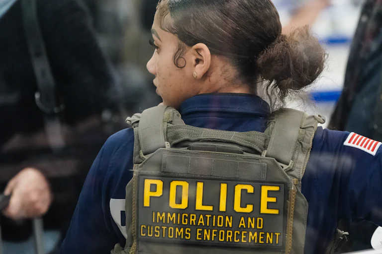 An ICE agent stands with her back to the camera and looks to the side