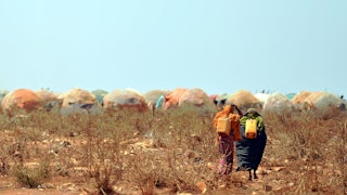 Two people walk away from the camera, surrounded by dried out vegetation, with tents in the background.