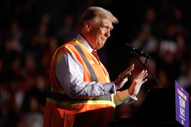 Donald Trump wears a garbage collector vest and raises both hands in front him