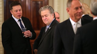 Elon Musk talks to Steve Bannon while he holds a glass in his hands.