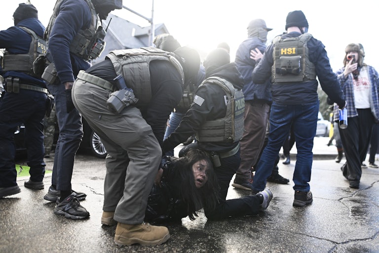Two ICE agents detain a woman on the paved road as other masked agents stand nearby.
