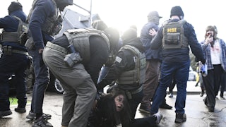 Two ICE agents detain a woman on the paved road as other masked agents stand nearby.