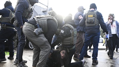 Two ICE agents detain a woman on the paved road as other masked agents stand nearby.
