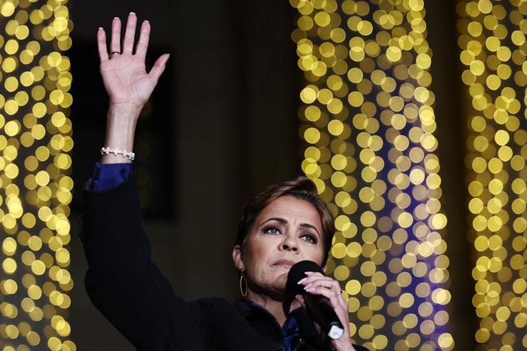 Kari Lake raises her hand while holding a microphone to her face