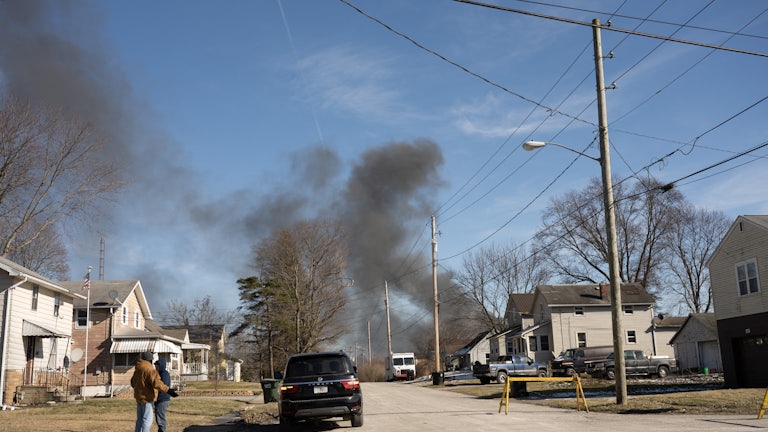 Smoke rises from a derailed cargo train in East Palestine, Ohio, in the background. Two people wearing coats stand in the left of the foreground, in a neighborhood.