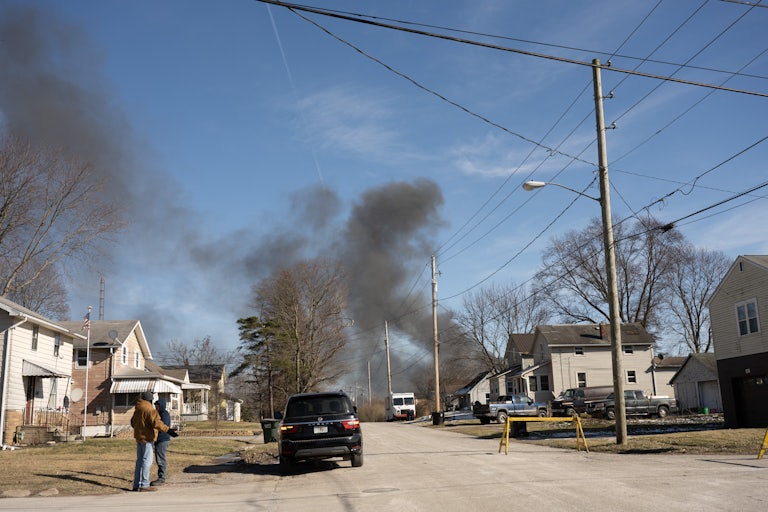 Smoke rises from a derailed cargo train in East Palestine, Ohio, in the background. Two people wearing coats stand in the left of the foreground, in a neighborhood.