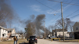 Smoke rises from a derailed cargo train in East Palestine, Ohio, in the background. Two people wearing coats stand in the left of the foreground, in a neighborhood.