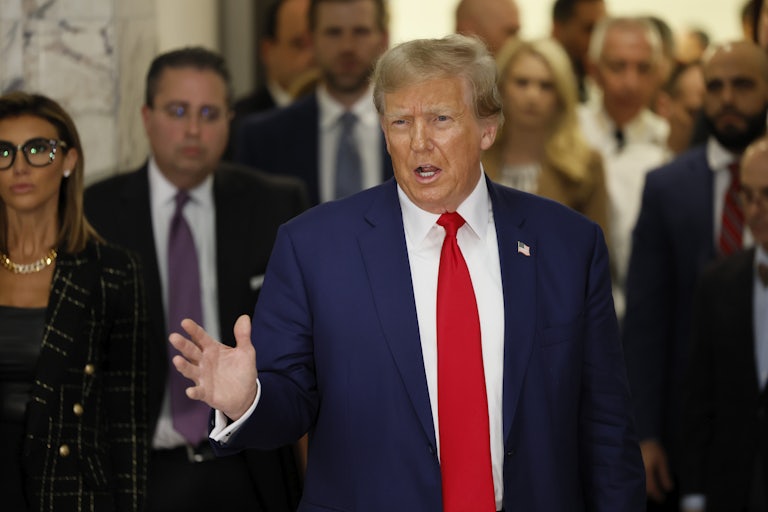 Donald Trump stands in the courtroom lobby saying something and gesturing with his hand. His lawyer Alina Habba stands to his left. Others are in the background.