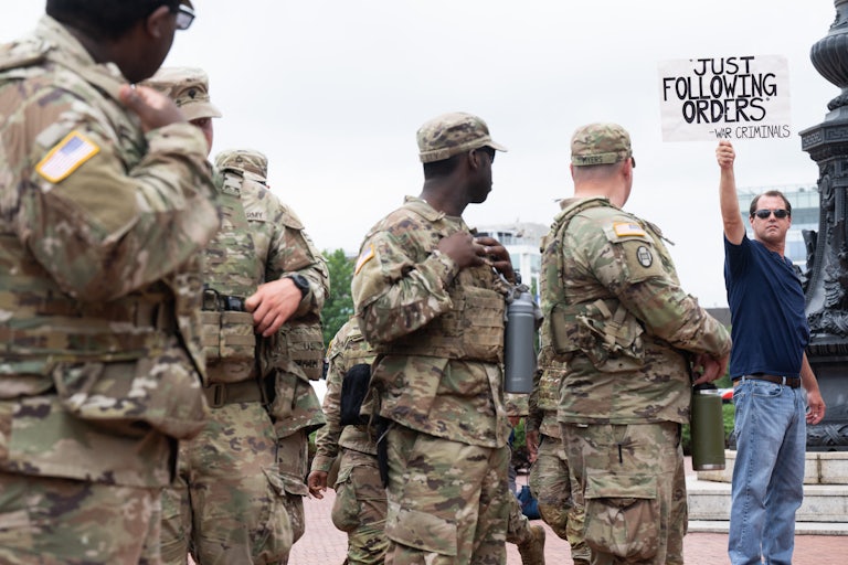National Guard members stand in Washington, D.C., next to a person holding up a sign that says, "'Just following orders' - war criminals"
