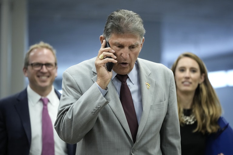 Senator Joe Manchin frowns while talking on his phone as two aides stand behind, smiling.