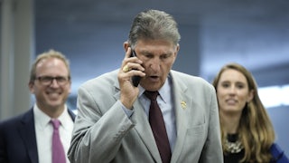 Senator Joe Manchin frowns while talking on his phone as two aides stand behind, smiling.