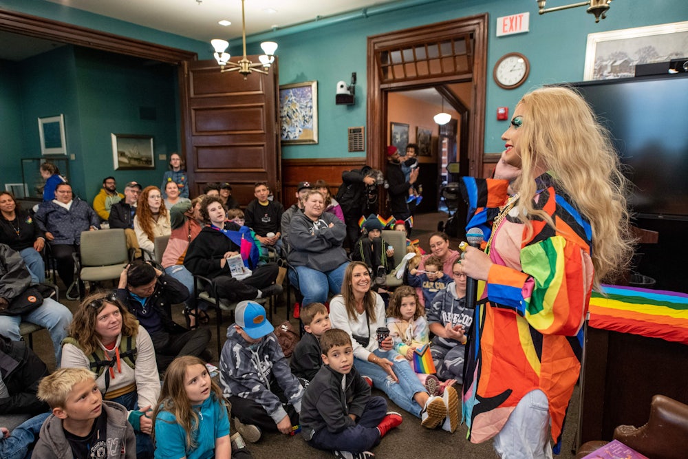 Children and adults sit and watch a drag performer standing with a microphone.