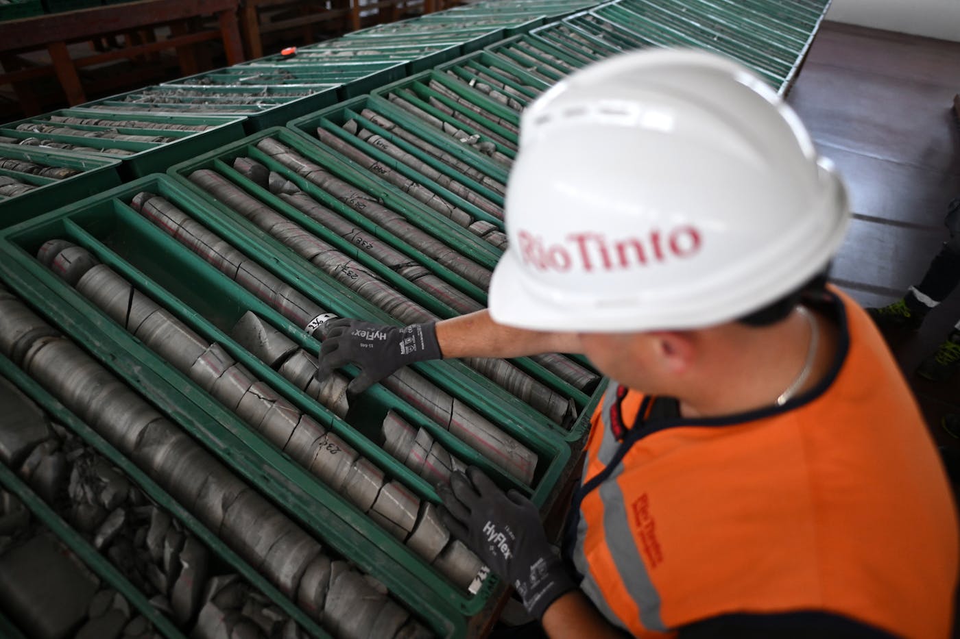 A person wearing a "Rio Tinto" helmet and orange vest examines trays of a grey mineral.