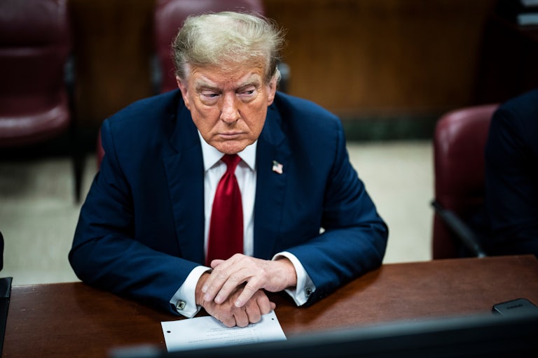 Donald Trump sits at a table with his hands folded on top while he looks to the side