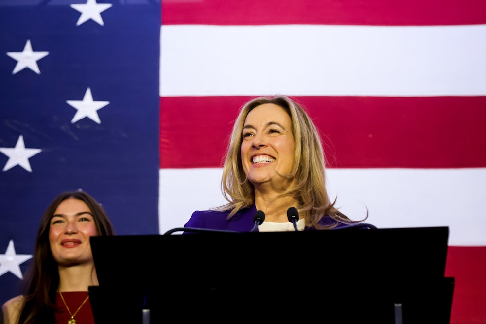 Mikie Sherill stands, smiling, at a podium in front of an American flag, with one person smiling in the background.