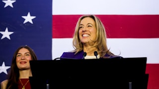 Mikie Sherill stands, smiling, at a podium in front of an American flag, with one person smiling in the background.