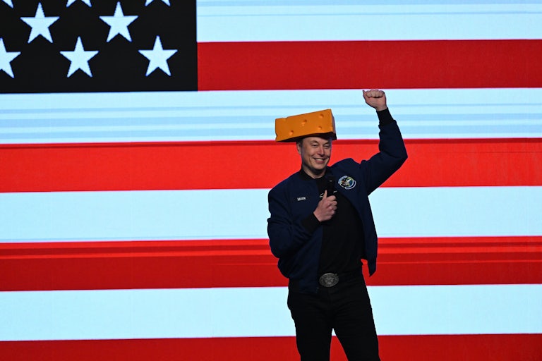 Elon Musk wears a cheese hat, holds a microphone, and raises his fist while on stage at a Wisconsin rally