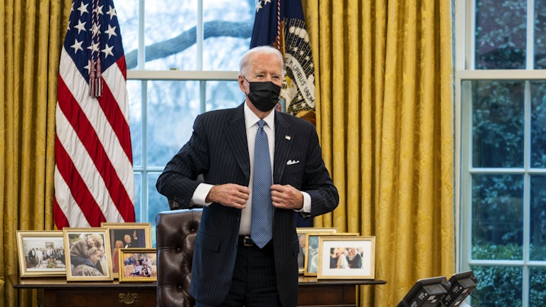 Joe Biden, wearing a surgical mask and standing in the Oval Office, adjusts his jacket after signing an executive order 