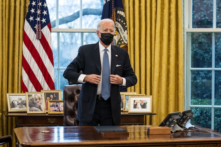 Joe Biden, wearing a surgical mask and standing in the Oval Office, adjusts his jacket after signing an executive order 