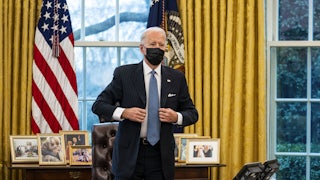 Joe Biden, wearing a surgical mask and standing in the Oval Office, adjusts his jacket after signing an executive order