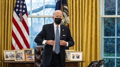 Joe Biden, wearing a surgical mask and standing in the Oval Office, adjusts his jacket after signing an executive order