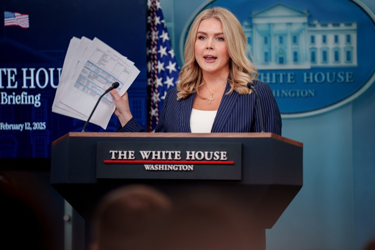 White House Press Secretary Karoline Leavitt holds multiple papers in the air as she speaks at the podium in the White House briefing room.