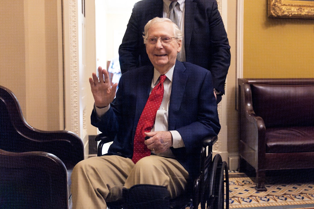 Senator Mitch McConnell, wearing a red tie and sitting in a wheelchair, heads to a vote. He is smiling and waving.