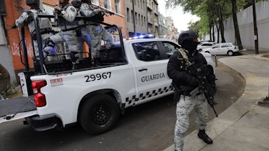 Armed members of the National Guard stand on the back of a pickup truck and on the sidewalk.