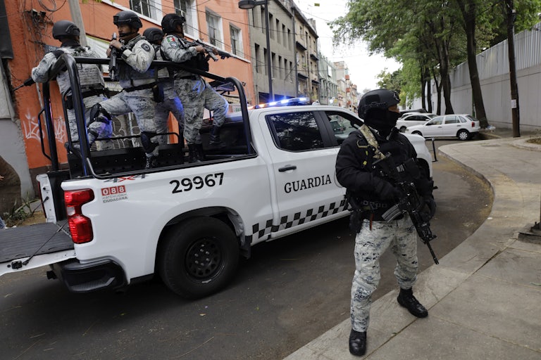 Armed members of the National Guard stand on the back of a pickup truck and on the sidewalk.