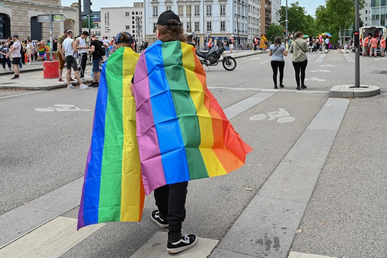 Two people with the LGBTQ Pride Flag draped ontheir backs walk in the street. Others with rainbow flags are also in the background.
