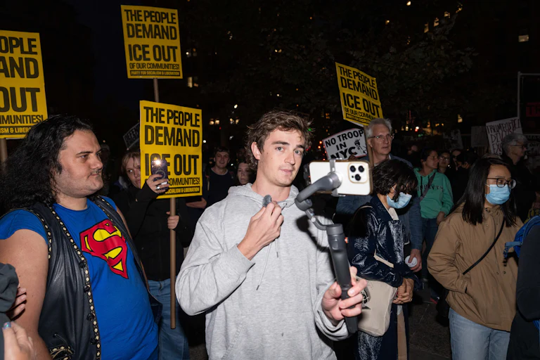 Nick Shirley holds an iPhone on a tripod while standing in the middle of an anti-ICE protest