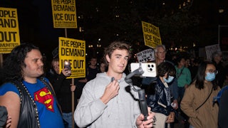 Nick Shirley holds an iPhone on a tripod while standing in the middle of an anti-ICE protest