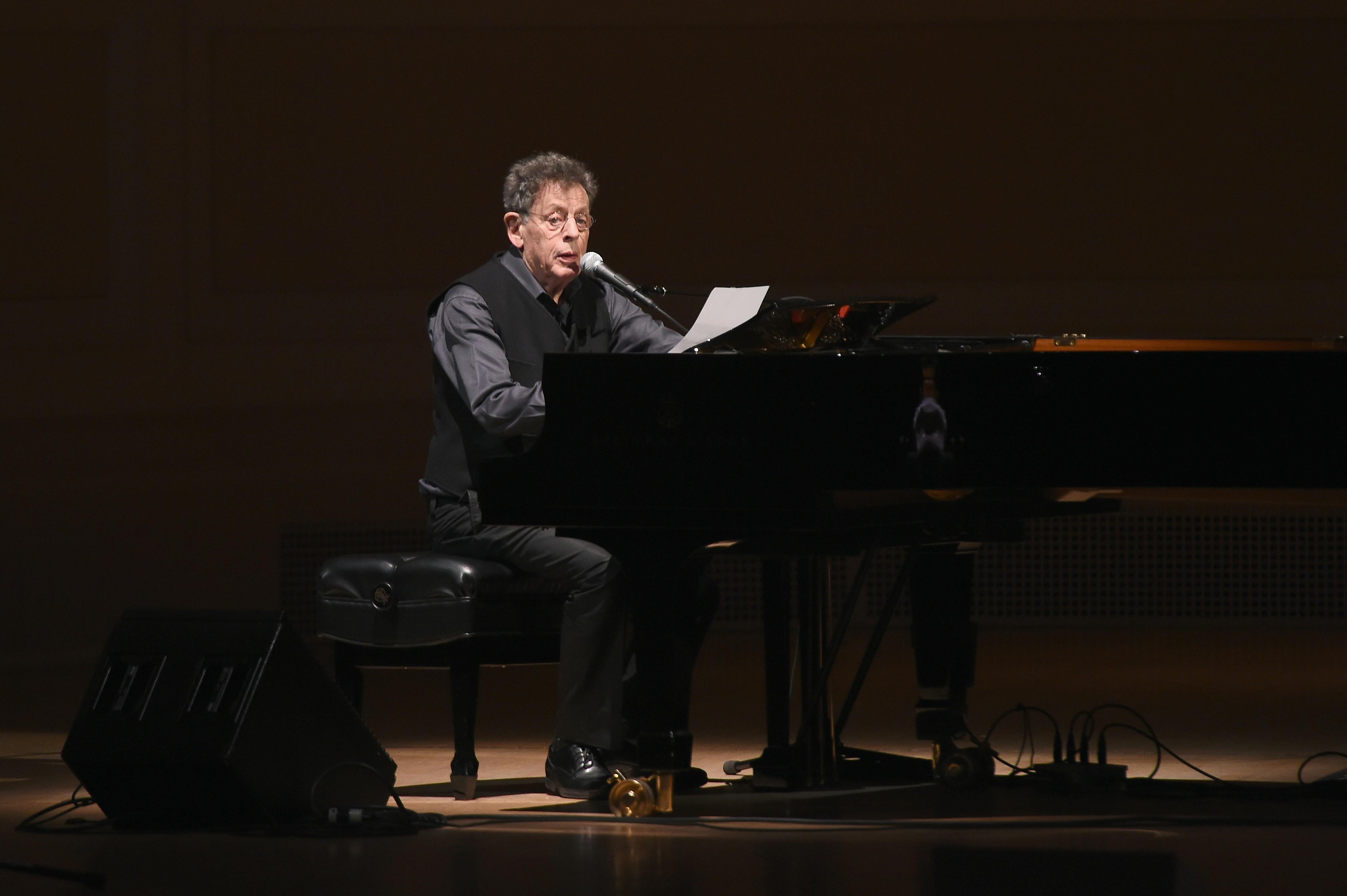Philip Glass reads into a mic while sitting at a piano on stage.