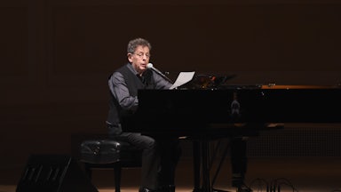 Philip Glass reads into a mic while sitting at a piano on stage.