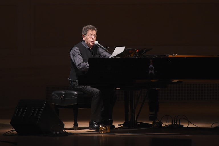 Philip Glass reads into a mic while sitting at a piano on stage.
