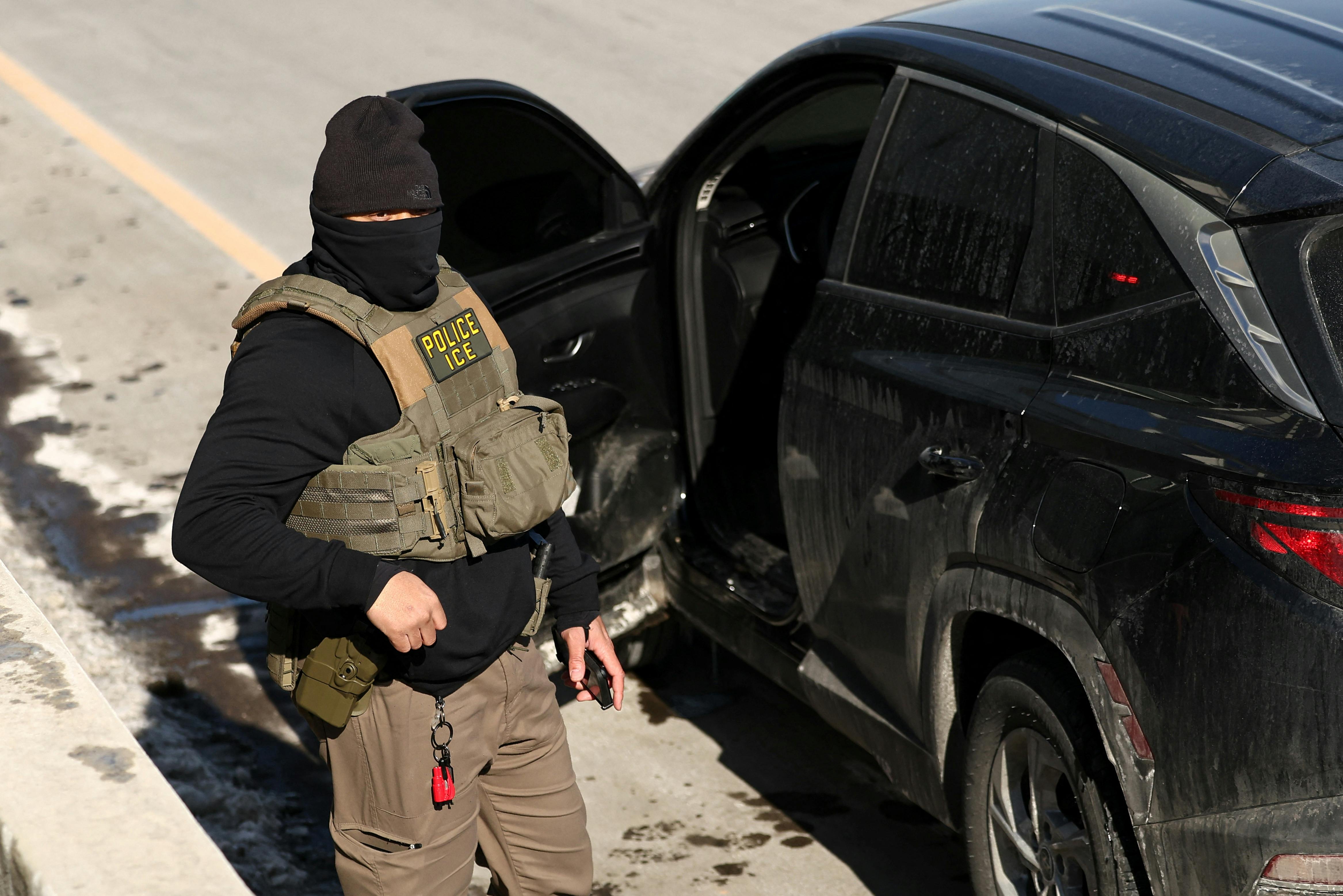 A masked ICE agent stands next to his car.