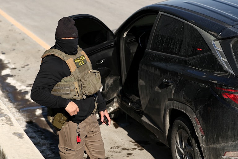 A masked ICE agent stands next to his car.