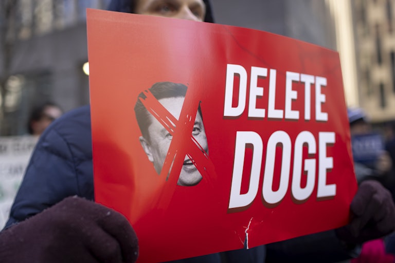 A protester holds up a sign that says, "Delete DOGE" next to a crossed-out photo of Elon Musk's face