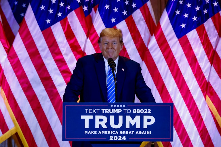 Donald Trump stands at a podium, in front of large American flags