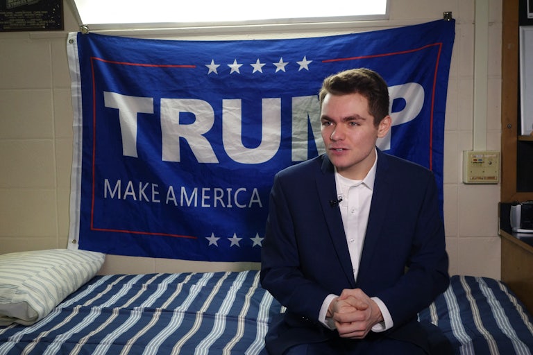 Nick Fuentes sits on a bed with a "Trump Make America Great Again" flag behind him