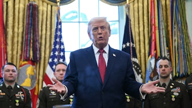Donald Trump holds his hands out to the side and speaks while standing at his desk in the Oval Office