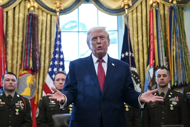 Donald Trump holds his hands out to the side and speaks while standing at his desk in the Oval Office