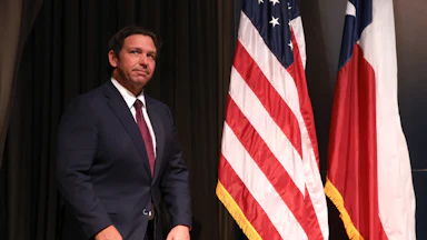 Florida Governor Ron DeSantis stands on stage next to the U.S. and Florida flags.