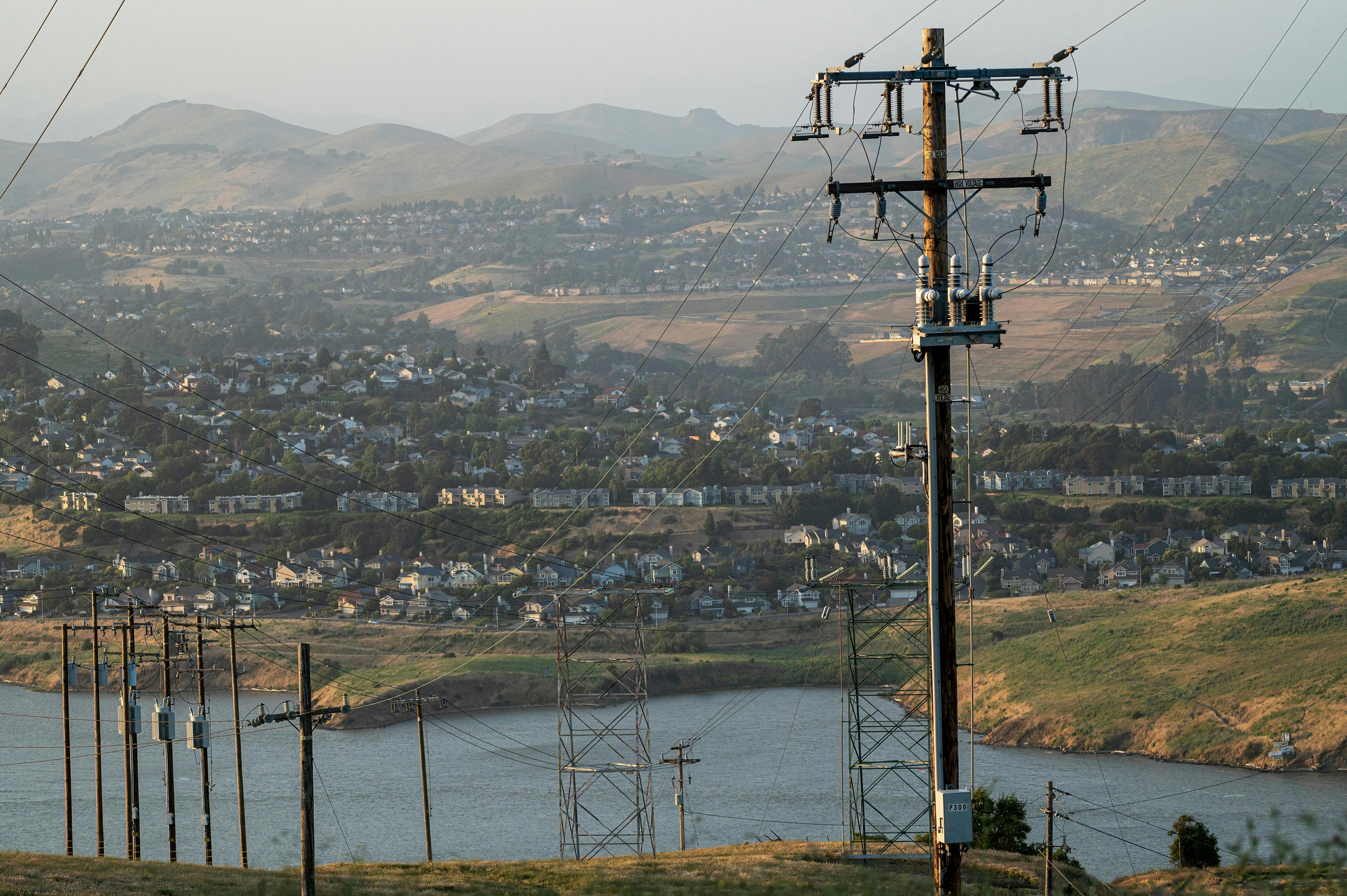 Power lines in the foreground are set against a neighborhood of homes on a hillside in the background. 