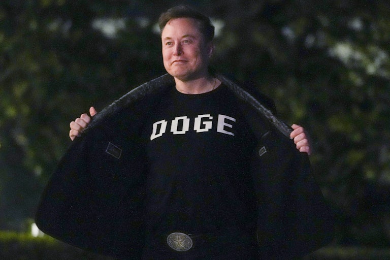 Elon Musk stands outside the White House and holds open his jacket to reveal the word "DOGE" on his shirt