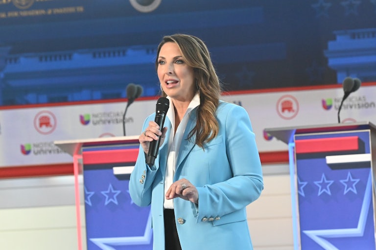 Ronna McDaniel at the Republican candidate debate hosted by Fox Business. She is on the stage with a mic in hand, wearing a light blue blazer. The candidate podiums with mics are behind her.
