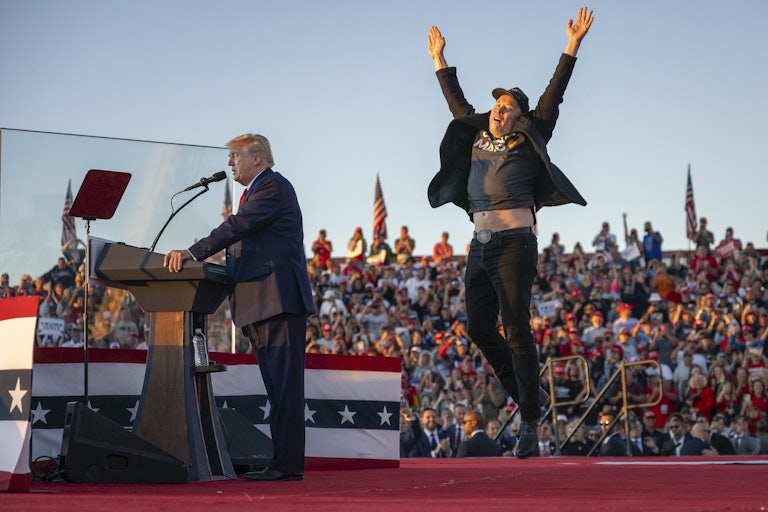 Elon Musk jumps in the air behind Donald Trump as he speaks at a lectern at his campaign rally